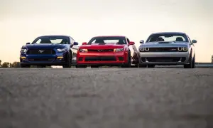 Trzy potężne muscle cary: niebieski Mustang, czerwony Camaro i srebrny Challenger, stoją obok siebie na tle zachodzącego słońca.