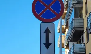 Znak zakaz postoju z tabliczką wskazującą, że zakaz obowiązuje na odcinku w obu kierunkach. Budynek z balkonami w tle.