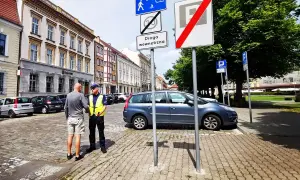 Policjant rozmawia z mężczyzną na brukowanej ulicy. Znaki drogowe wskazują strefę zamieszkania i koniec strefy parkowania.