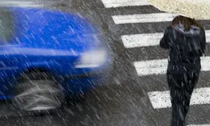 Deszczowa pogoda na przejściu dla pieszych. Niebieski samochód mija kobietę z parasolem.