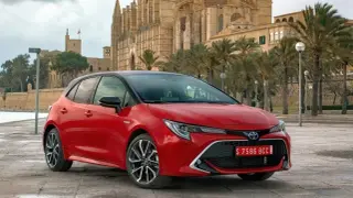 Czerwona Toyota Corolla E21 z czarnym dachem zaparkowana przed katedrą i palmami.