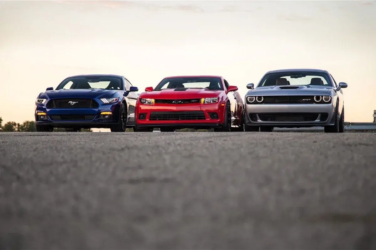 Trzy potężne muscle cary: niebieski Mustang, czerwony Camaro i srebrny Challenger, stoją obok siebie na tle zachodzącego słońca.
