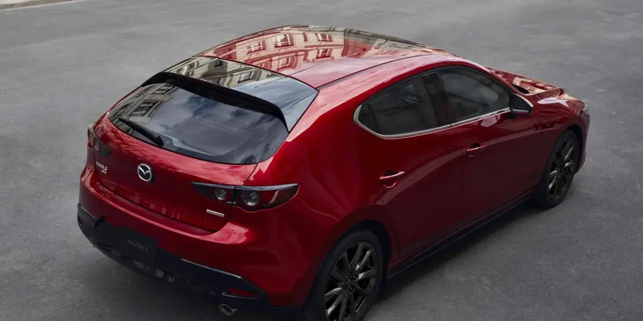 Czerwony hatchback Mazda 3 z czarnymi felgami, odbijający budynek na dachu. Wygląda elegancko, w przeciwieństwie do nudnego sedana.