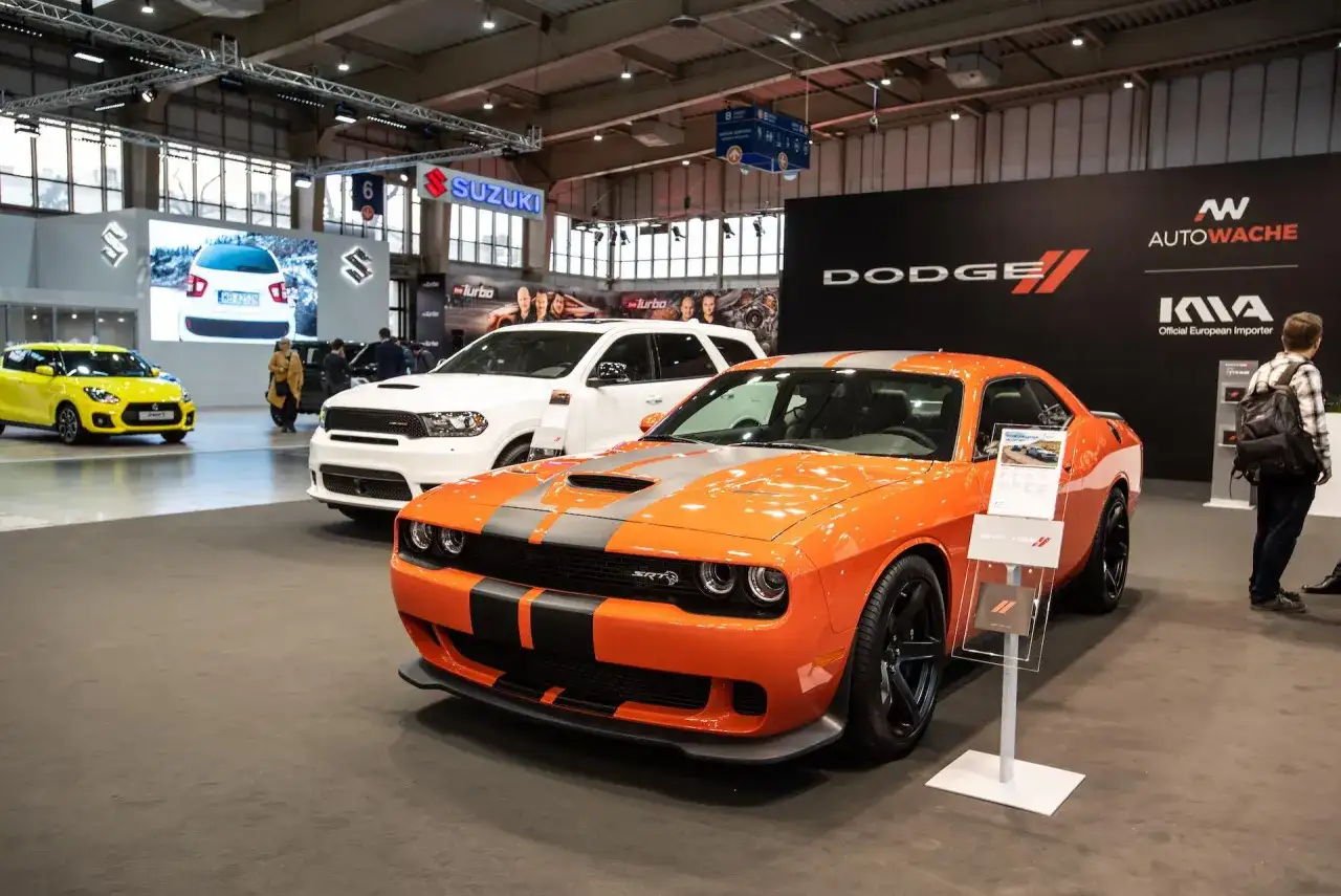Pomarańczowy Dodge Challenger SRT na wystawie. Sprawdź aktualną dodge challenger cena i porównaj z białym SUV-em w tle.
