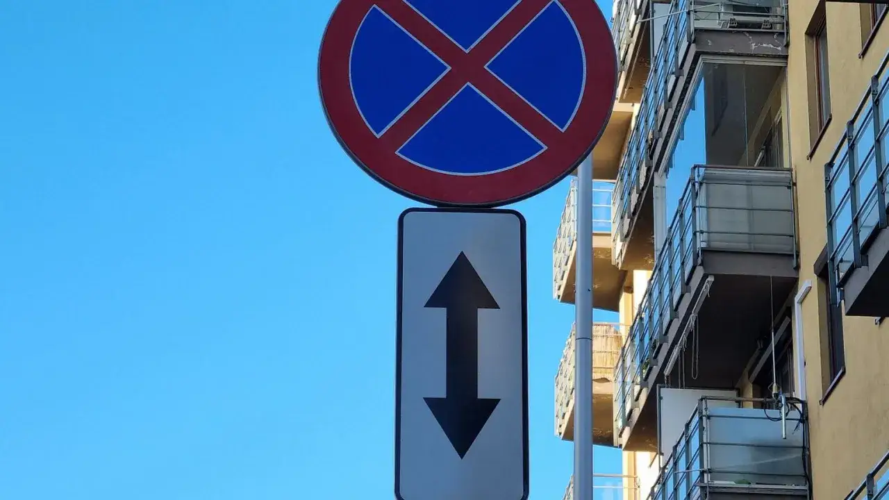 Znak zakaz postoju z tabliczką wskazującą, że zakaz obowiązuje na odcinku w obu kierunkach. Budynek z balkonami w tle.