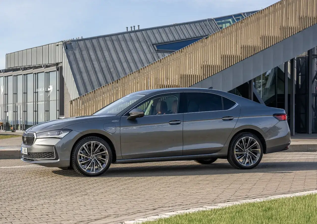 Elegancka, szara Skoda Superb 2024 z nowoczesnymi felgami stoi przed budynkiem z przeszkloną fasadą.