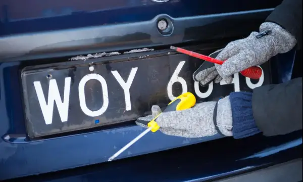 Ręce w rękawiczkach odkręcają śruby tablicy rejestracyjnej.