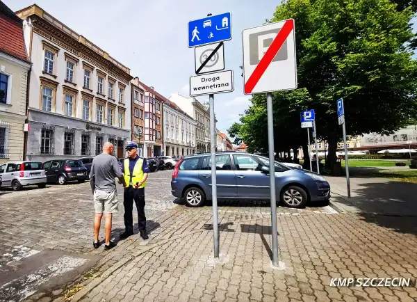 Policjant rozmawia z mężczyzną na brukowanej ulicy. Znaki drogowe wskazują strefę zamieszkania i koniec strefy parkowania.