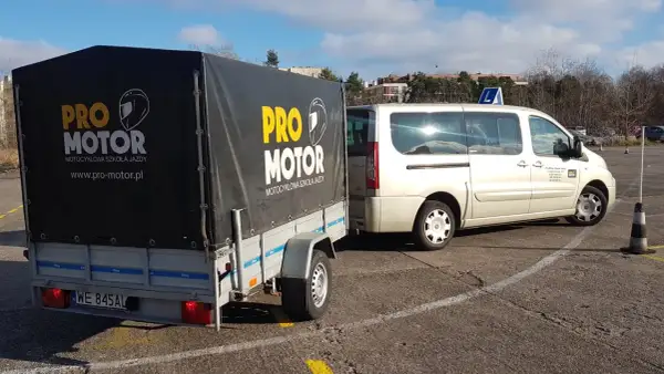 Przyczepa szkoły jazdy b+e "Pro Motor" z motocyklem w środku, obok biały bus.