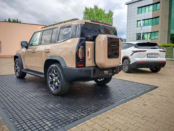 Beżowy Jetour T2 z kołem zapasowym z tyłu, obok białego SUV-a. Sprawdź Jetour T2 cena i dostępne opcje.