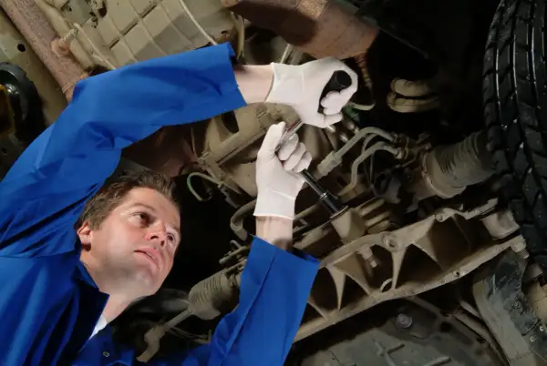 Mechanik w niebieskim kombinezonie naprawia samochód, używając klucza do dokręcenia śruby. Jego biała manszeta jest widoczna.
